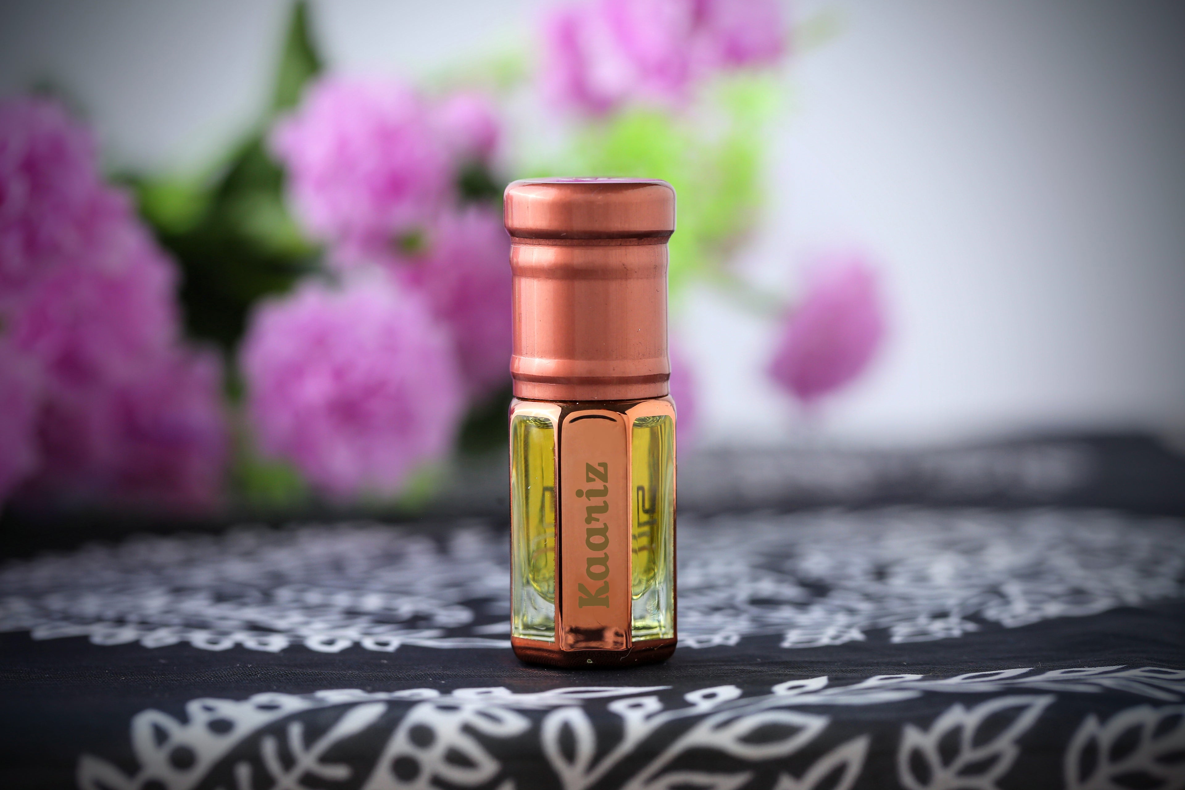 Kaariz Attar by ARK Fragrances in rose gold bottle. Premium floral long-lasting perfume oil displayed with soft purple flowers. Best sweet alcohol-free scent in Pakistan.